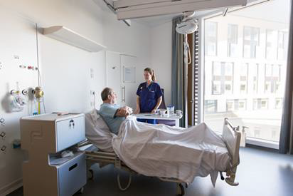 The nurse is treating a patient whilst in the bay bed in a well-lit room.