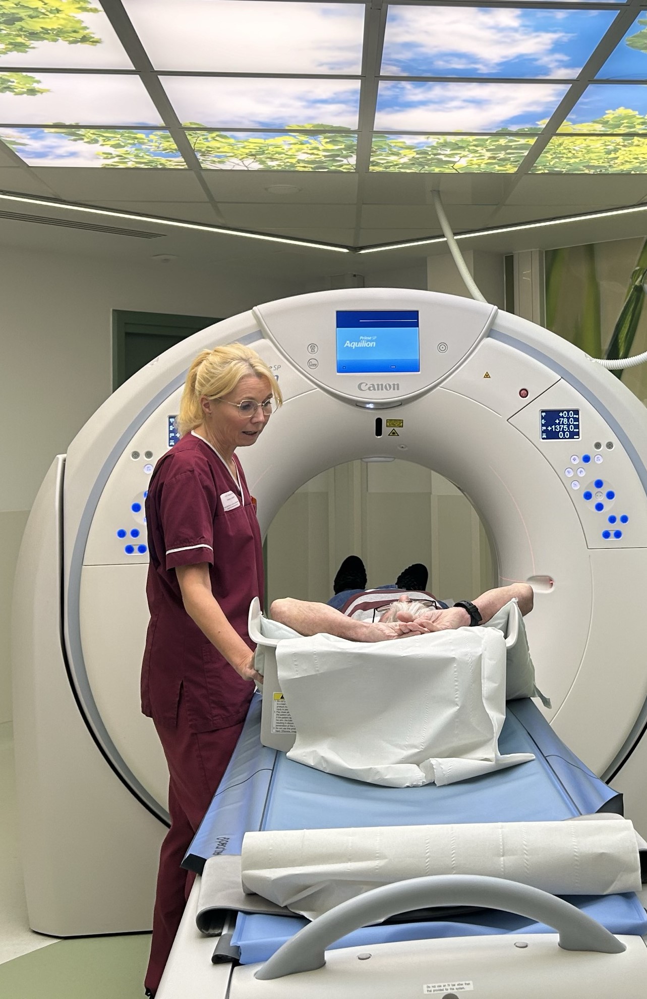 A radiographer helps a patient into the CT scanning machine. Above them is a light box with nature-inspired artwork 